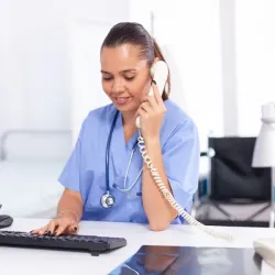 Female medical professional wearing blue scrubs and a stethoscope, smiling while answering a telephone call at a desk with a computer keyboard