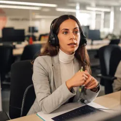 Female customer service representative wearing a white turtleneck and grey blazer, listening attentively with a headset while holding a pen at her desk in an office setting