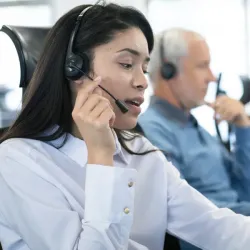 Customer service representative focused on a call, wearing a white button-up shirt and headset, with another team member visible in the background