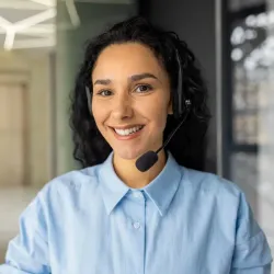 Female customer service representative with curly dark hair wearing a light blue button-up shirt and headset, smiling warmly at the camera